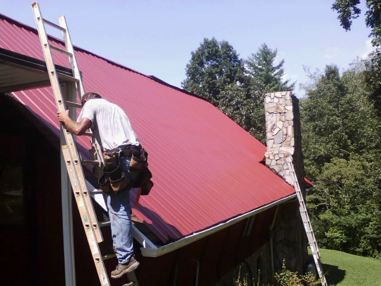 Roofer on a ladder beside a red metal roof and stone chimney — Soffit & Fascia Repair by Roofing Brothers in Exmore